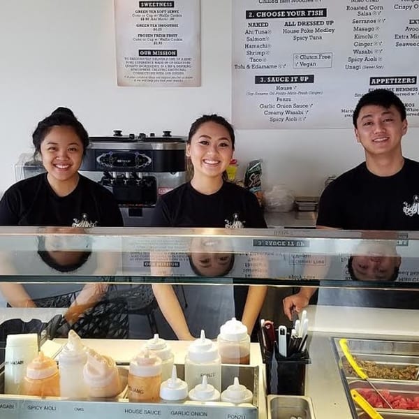 Three smiling Poke Poke Fish Bar employees stand behind a counter with sauces and poke ingredients.