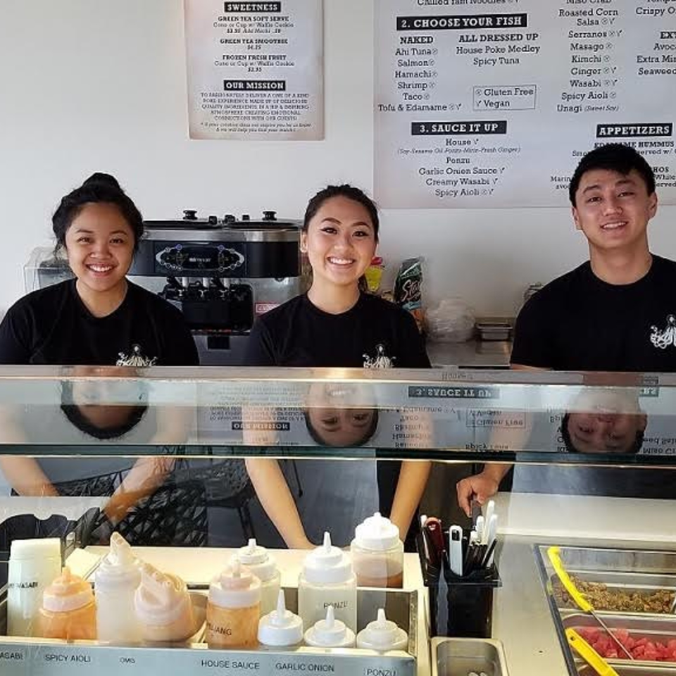 Three smiling Poke Poke Fish Bar employees stand behind a counter with sauces and poke ingredients.