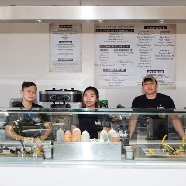 Three Poke Poke Fish Bar employees stand behind a counter with menu boards above them.