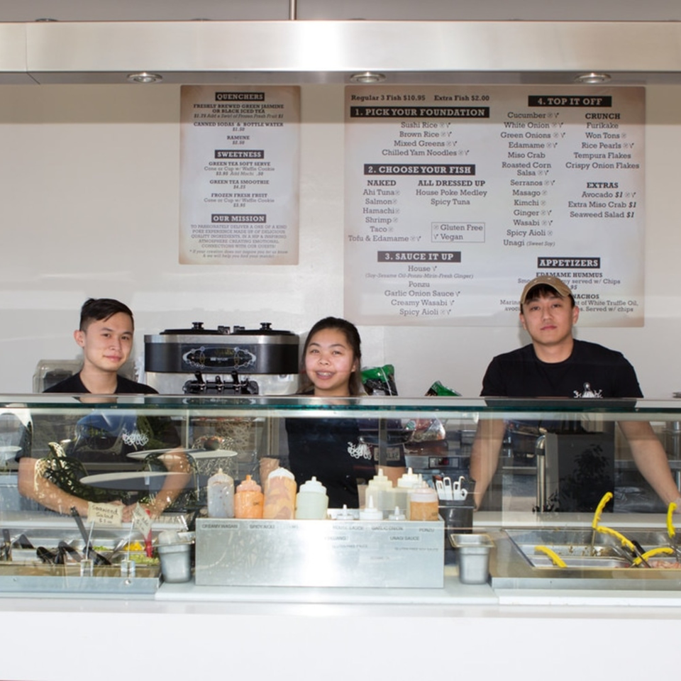 Three Poke Poke Fish Bar employees stand behind a counter with menu boards above them.