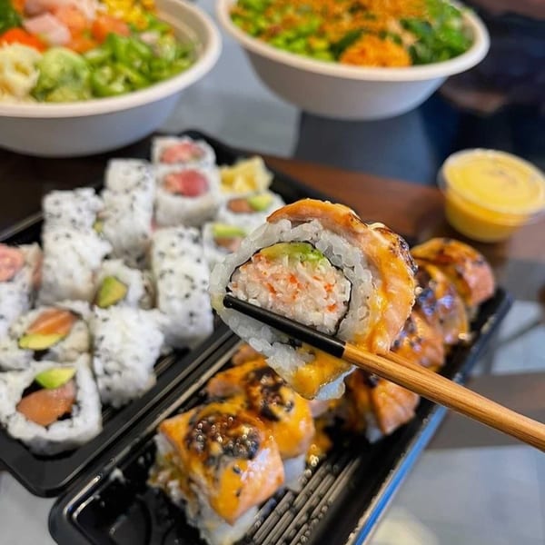 Chopsticks holding a sushi roll with crab and avocado, with more sushi and poke bowls in the background.