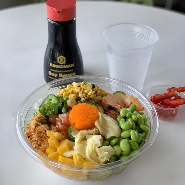 A colorful poke bowl with salmon, edamame, corn, mango, and roe, with soy sauce and hot chips.
