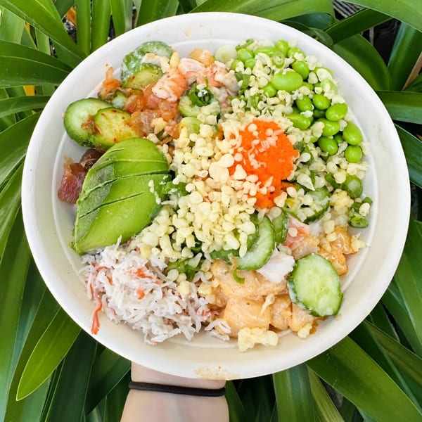 A vibrant poke bowl with avocado, edamame, shrimp, cucumber, and orange roe, held against green leaves.