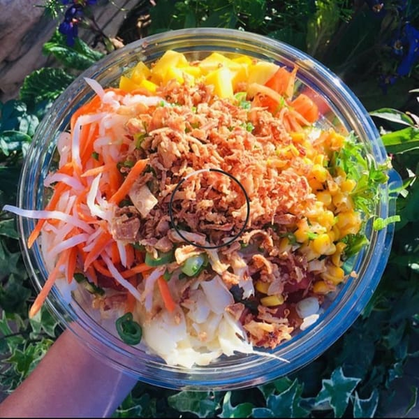 Poke bowl with tuna, pineapple, corn, shredded carrots, and crispy onions, held outdoors.