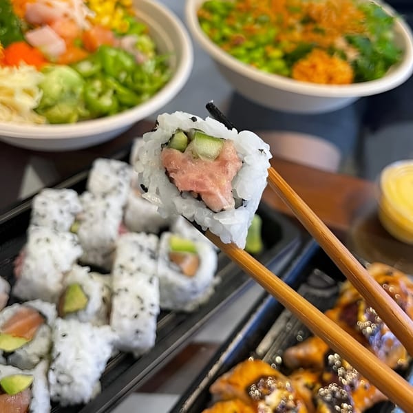 Chopsticks holding a tuna and cucumber sushi roll, with poke bowls and more sushi in the background.