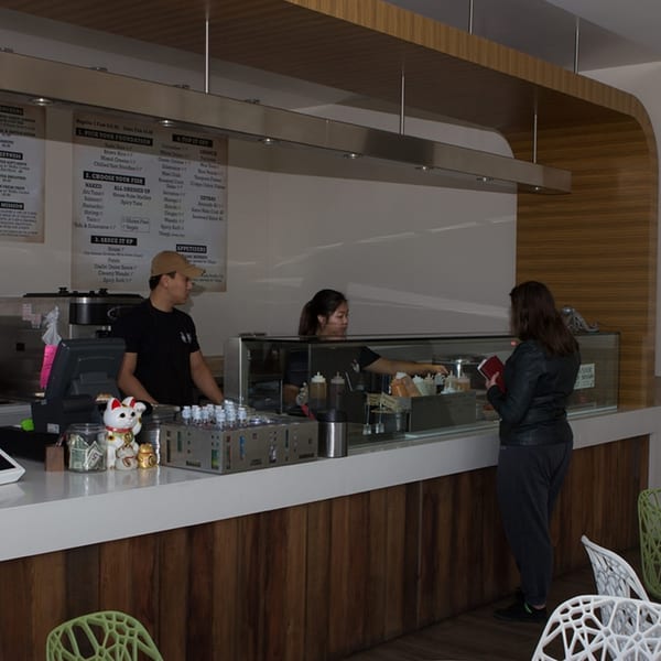 Two Poke Poke Fish Bar employees serve a customer at the counter with menu boards overhead.
