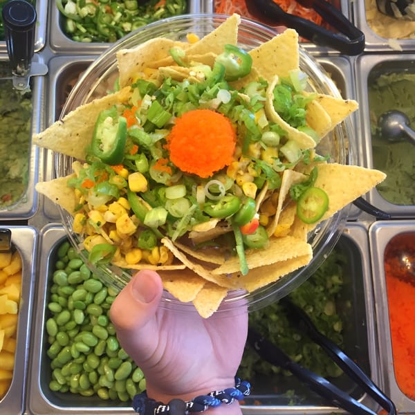 A hand holds a Poke Poke Fish Bar poke bowl with tortilla chips, corn, jalapeños, and orange roe.