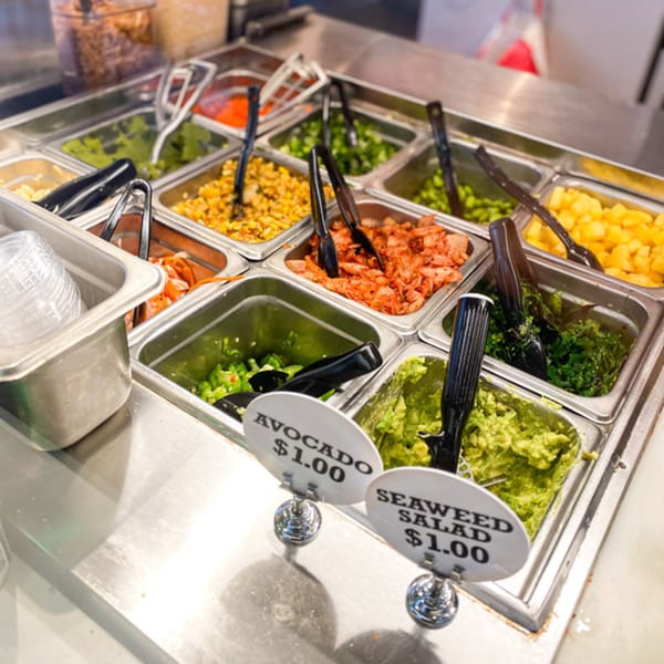 A poke bar with various toppings in metal bins, including avocado, seaweed salad, corn, and kimchi.