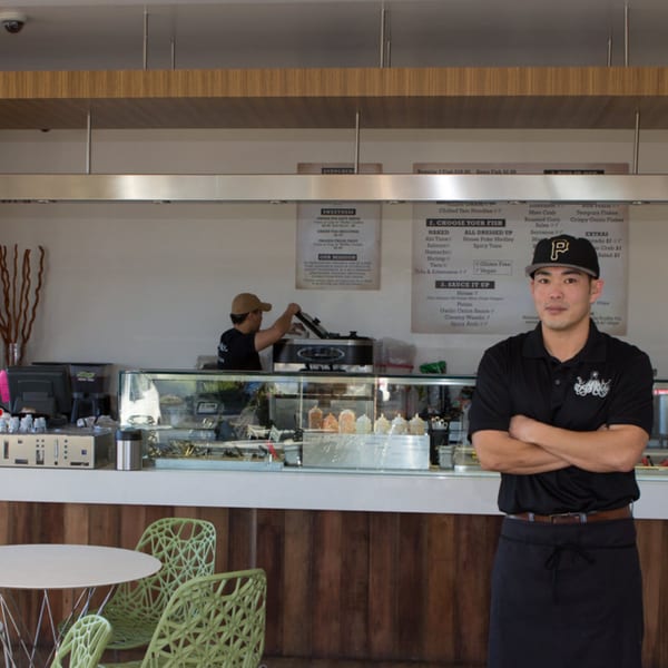 A Poke Poke Fish Bar employee in a black hat and shirt stands in front of the counter.