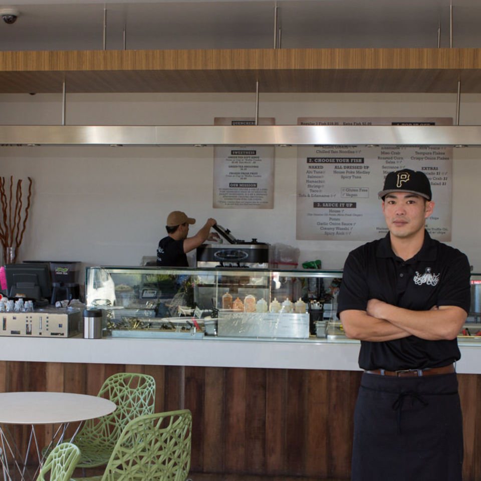 A Poke Poke Fish Bar employee in a black hat and shirt stands in front of the counter.